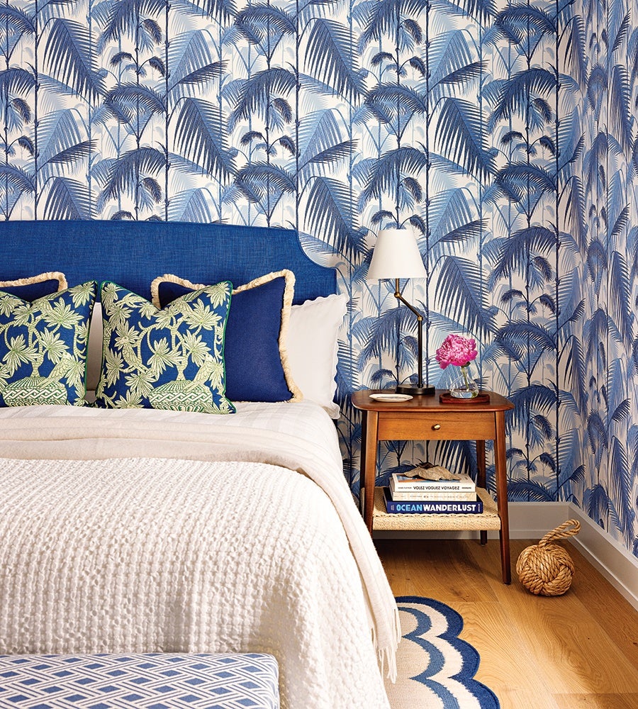 A bedroom corner featuring a bed with a blue upholstered headboard and white bedding. The bed has decorative pillows with green palm leaf patterns and solid blue pillows with beige trim. The wall behind the bed is covered in blue and white palm leaf patterned wallpaper. Next to the bed is a wooden nightstand with a drawer, holding a modern black and brass table lamp with a white shade, a small pink flower in a glass vase, and a few books on the lower shelf. The floor is light wood with a blue and white scalloped rug partially visible. A woven rope decorative object sits on the floor near the nightstand.