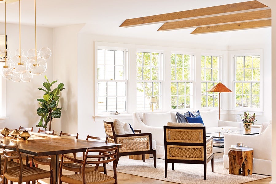 Bright living and dining area with large windows, featuring a wooden dining table with woven chairs, a white sectional sofa, two woven armchairs, a wooden side table, a floor lamp with an orange shade, and a cluster of glass pendant lights. The room has white walls, wooden ceiling beams, and a potted plant in the corner.