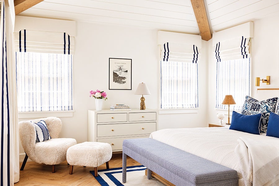 Bedroom with white walls and wooden ceiling beams, featuring a white dresser with gold knobs, a white lamp, and a vase of pink flowers. There is a cozy cream-colored chair and matching ottoman with blue and white striped pillows. The bed has white bedding with blue and patterned pillows, and a light blue bench at the foot. Windows have white Roman shades with navy blue stripes, and a blue and white rug is partially visible on the wooden floor.