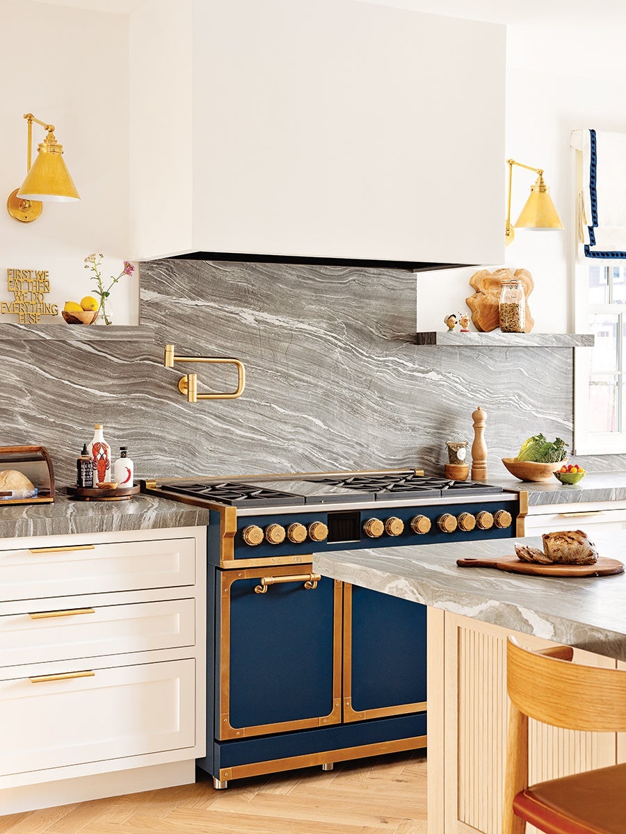 A modern kitchen corner featuring a navy blue and gold-accented stove with multiple knobs and a large cooktop. The backsplash and countertops are made of gray marble with white veining. White cabinetry with gold handles is visible on the left, and a wooden chair with a red cushion is partially visible on the right. The kitchen has gold wall-mounted lamps, a pot filler faucet above the stove, and various kitchen items including a loaf of bread on a wooden cutting board, a bowl of vegetables, and jars on the countertop. The overall design combines elegant materials with warm wood tones.