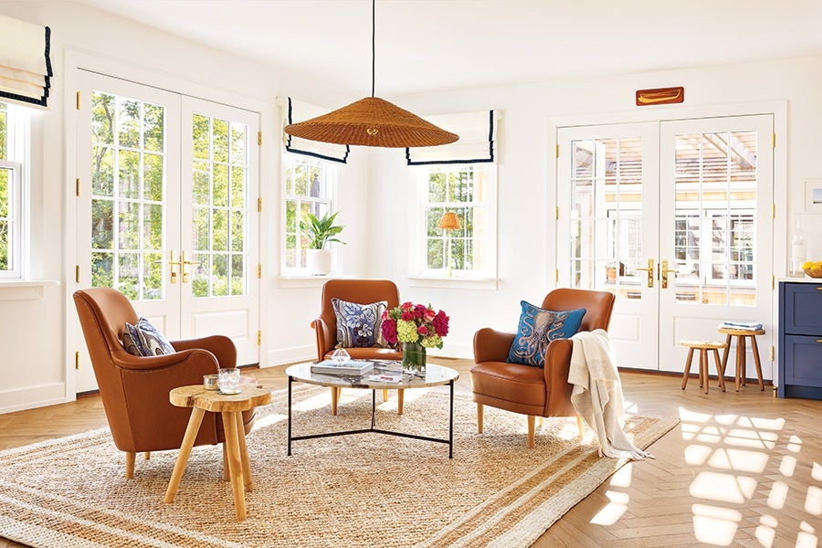 Bright living room with three brown leather armchairs, each adorned with decorative pillows. A round glass coffee table with a black metal frame sits in the center, holding books and a vase of colorful flowers. A small wooden side table with a candle is next to one armchair. The room features large windows and French doors letting in natural light, white walls, a woven area rug, and a large woven pendant light hanging from the ceiling.