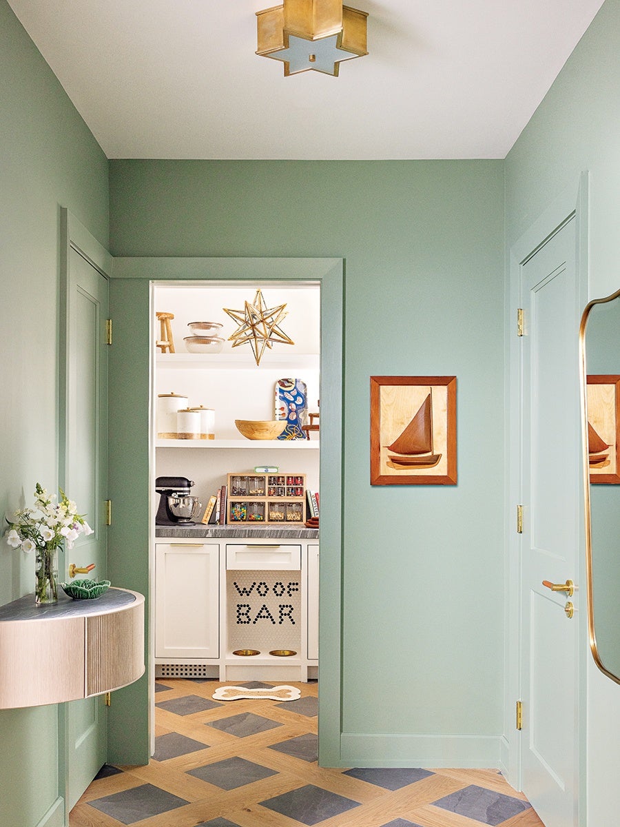 Hallway with sage green walls and two closed doors, featuring a small wall-mounted console table with a vase of white flowers and a green decorative dish. The floor has a geometric pattern with wood and dark tiles. A wooden framed artwork of a sailboat hangs on the right wall. Through an open doorway, a bright pantry or kitchen area is visible with white cabinetry, a black mixer, and a pet feeding station labeled "WOOF BAR" with two gold bowls and a bone-shaped rug. A star-shaped pendant light hangs in the pantry, and a gold star-shaped ceiling light fixture is mounted in the hallway. A tall mirror with a gold frame is partially visible on the right.