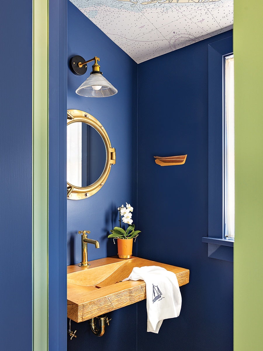 A small bathroom corner with deep blue walls and a nautical theme. It features a wooden sink with a brass faucet, a round brass porthole-style mirror, and a clear glass wall sconce with brass accents. A white towel with a sailboat embroidery is draped over the sink, and a small potted orchid sits on the sink countertop. The ceiling is decorated with a nautical map, and a small wooden boat shelf is mounted on the wall. A window with a blue frame lets in natural light.