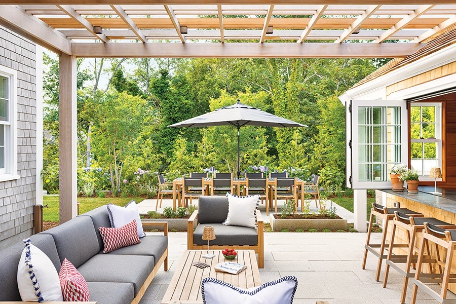 Outdoor patio with a wooden pergola overhead, featuring a gray cushioned sofa and armchair with decorative pillows, a wooden coffee table, and a dining area in the background with a large umbrella. There is also a bar counter with wooden stools on the right side, surrounded by greenery and trees.