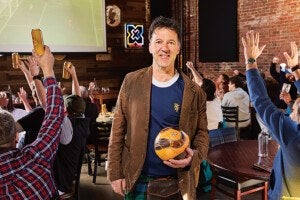 A man wearing a brown blazer over a blue shirt and a green kilt is standing indoors, holding a yellow and black soccer ball. Behind him, people are seated at tables, raising their hands and holding drinks, watching a soccer game projected on a screen. The setting has exposed brick walls and a casual, lively atmosphere.