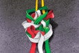 A colorful abstract sculpture composed of intertwined, ribbon-like shapes in red, green, and white, suspended by two yellow straps against a textured gray background.