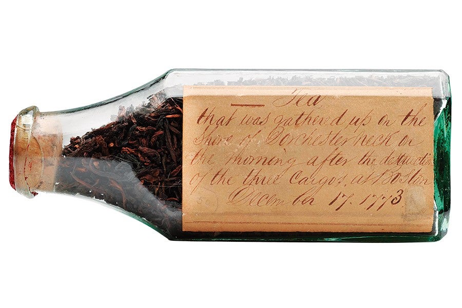 A small glass bottle sealed with a cork, containing loose black tea leaves. The bottle has an old, handwritten label that reads: "Tea that was gathered up on the shore of Dorchester neck on the morning after the destruction of the three Cargos, at Boston December 17, 1773.