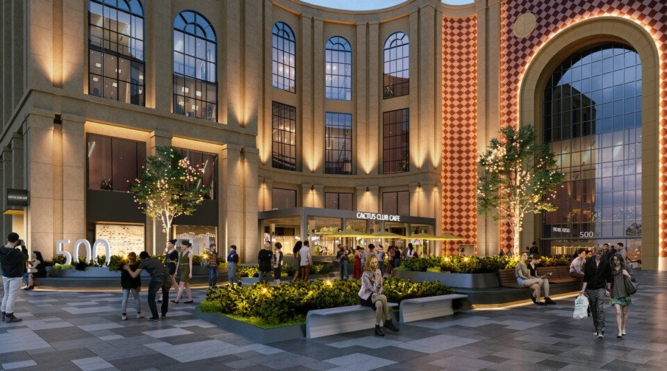 Outdoor plaza area in front of a building with large arched windows and warm lighting. The building has a sign for "Cactus Club Cafe." The plaza features modern benches, planters with greenery, and illuminated trees. People are walking, sitting, and socializing throughout the space. The ground is paved with large, square tiles in varying shades of gray. A large "500" sign is visible near the left side of the image.