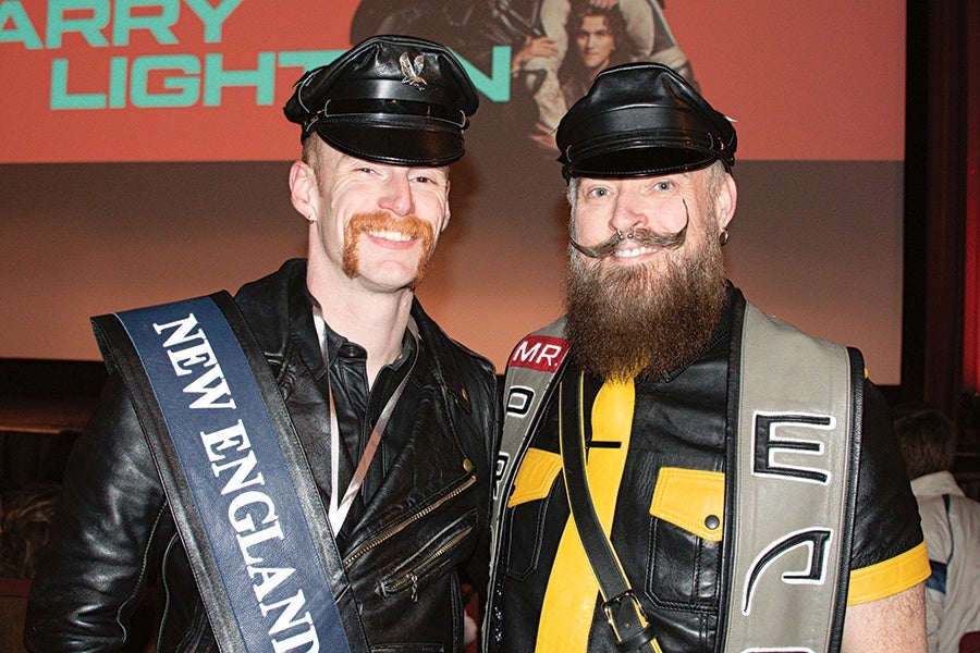 Two men wearing black leather caps and jackets stand side by side. The man on the left has a red mustache and wears a sash that reads "NEW ENGLAND." The man on the right has a full beard, curled mustache, and a sash with the letters "E" and "J" visible. Both are smiling.
