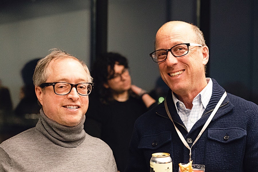 Two middle-aged men wearing glasses are smiling. The man on the left is wearing a gray turtleneck sweater, and the man on the right is wearing a navy blue jacket over a light blue collared shirt, holding a can and a sandwich. A blurred person is visible in the background.