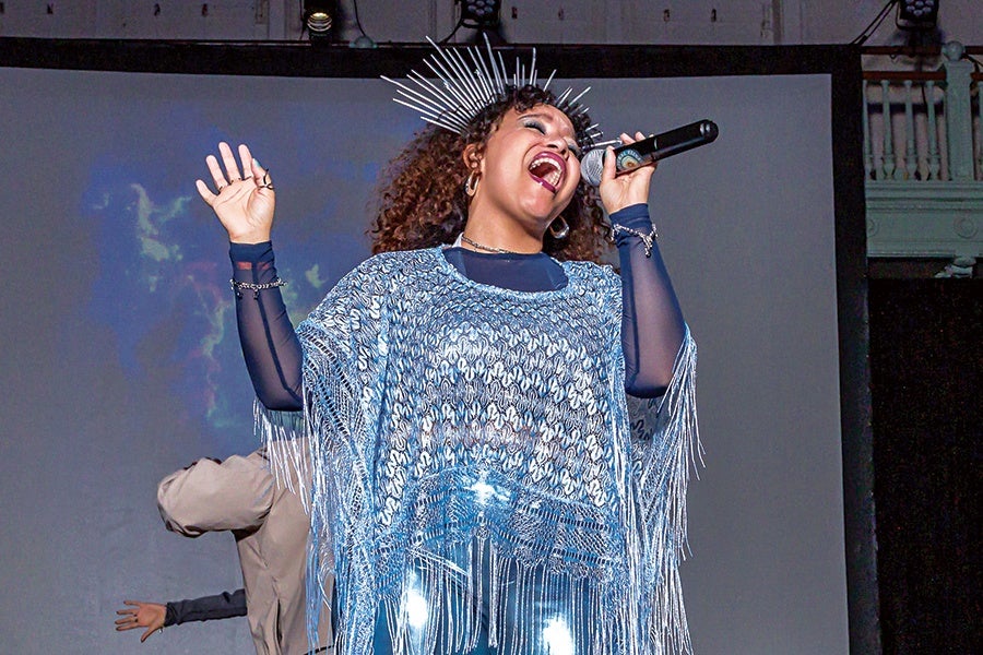 A woman passionately singing into a microphone, wearing a shiny silver fringed poncho over a dark long-sleeve top. She has curly hair and is adorned with a spiked silver headpiece and hoop earrings. Her eyes are closed, and her mouth is open wide as she performs. The background is dark with a large screen behind her.