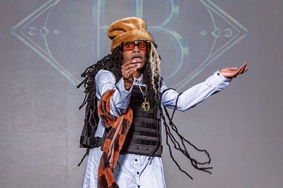 Person with long dreadlocks wearing a tan fur hat, orange-tinted glasses, a light blue button-up shirt, a black tactical vest, and a brown and orange patterned scarf, gesturing with both hands against a gray background with a faint geometric design.