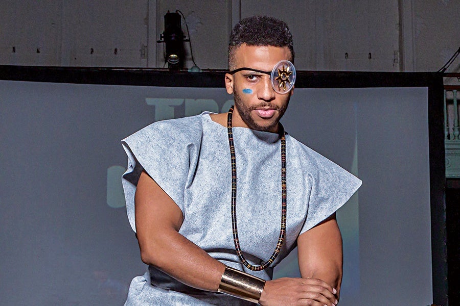 A man wearing a light gray, sleeveless tunic with wide shoulders, a long beaded necklace, and a metallic cuff bracelet on his right wrist. He has a decorative eyepiece with a sun design over his left eye and a blue paint mark on his right cheek. His hair is short and curly.