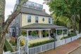 Two-story house with yellow siding on the front and gray shingles on the side, featuring a large covered porch with white columns and hanging ferns. The porch has seating with cushions, and the house has a white picket fence surrounding the front yard. A brick sidewalk runs alongside the fence, and large trees provide shade around the property.