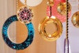 Decorative hanging ornaments featuring circular designs, including a large ring filled with small blue and green beads, a gold ring with a smooth reflective surface, and a smaller circular piece adorned with purple, white, and black beads arranged in a radial pattern. The ornaments are suspended by strings of gold and black beads against a wooden background.
