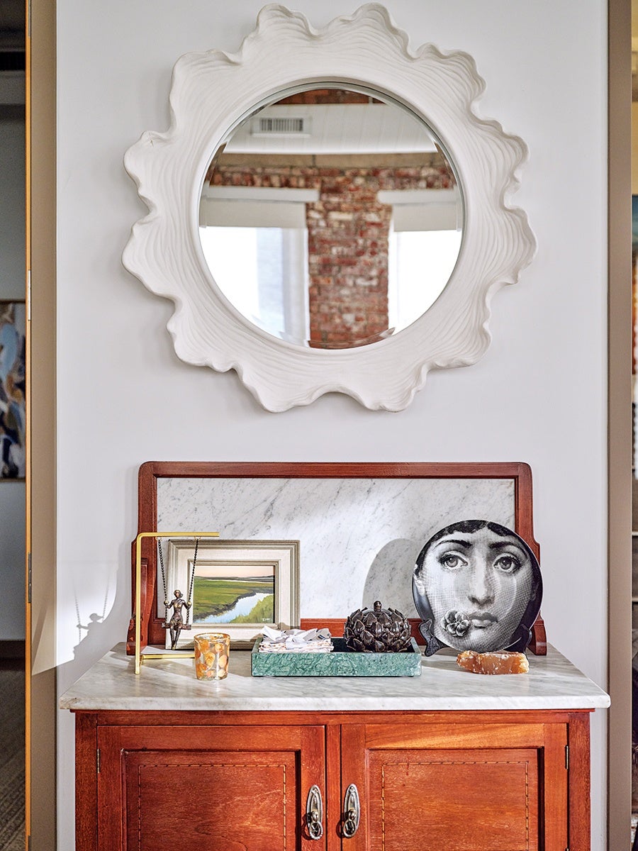 A wooden cabinet with a white marble top is shown against a white wall. Above the cabinet hangs a round mirror with a wavy, textured white frame. On the cabinet's surface, there is a small framed landscape painting, a brass swing sculpture with a small figure, a candle with an orange pattern, a green marble tray holding papers, a dark decorative object resembling a pinecone, a black and white plate featuring a woman's face with a flower in her mouth, and a piece of orange crystal or mineral. A rectangular marble slab with a wooden frame leans against the wall behind the items.