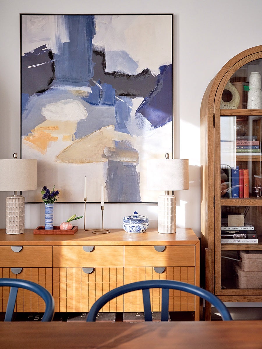 Wooden sideboard with a large abstract painting in shades of blue, white, and beige above it. On the sideboard are two beige table lamps with textured bases, a blue and white ceramic jar, a small vase with purple flowers, a pink decorative object, and a minimalist gold candle holder with two white candles. To the right is a wooden cabinet with glass doors containing books and decorative items. The foreground shows the backs of two blue chairs.