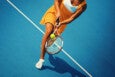 A female tennis player wearing an orange dress and white sneakers is hitting a tennis ball with a racket on a blue tennis court. The image captures her mid-action near a white boundary line.