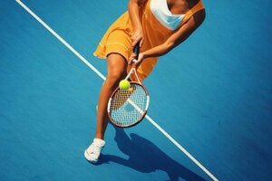 A female tennis player wearing an orange dress and white sneakers is hitting a tennis ball with a racket on a blue tennis court. The image captures her mid-action near a white boundary line.