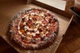 A pizza in a cardboard box with a thick crust, topped with tomato sauce, melted cheese, dollops of white sauce, and scattered pieces of browned sausage. Grated cheese is generously sprinkled around the crust and inside the box. The pizza is on a wooden surface with a plastic container partially visible to the side.