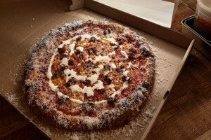 A pizza in a cardboard box with a thick crust, topped with tomato sauce, melted cheese, dollops of white sauce, and scattered pieces of browned sausage. Grated cheese is generously sprinkled around the crust and inside the box. The pizza is on a wooden surface with a plastic container partially visible to the side.