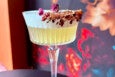 A pale yellow cocktail in a clear, fluted glass with a stem. The rim is coated with crushed red and brown spices and garnished with small dried rosebuds. The background features a blurred floral pattern with red, orange, and blue hues.
