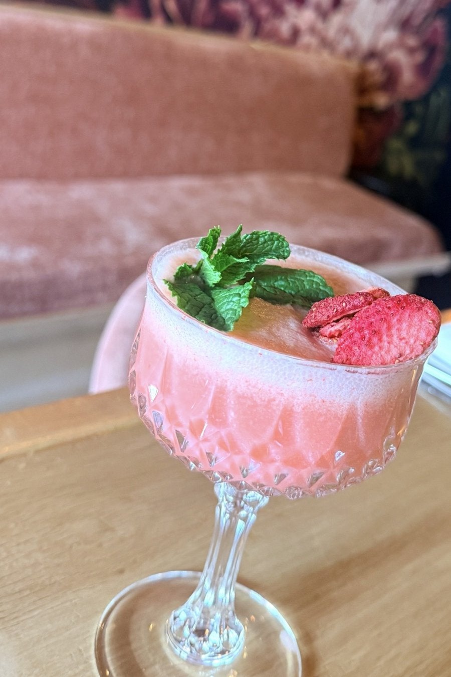 Pink frothy cocktail served in a decorative crystal coupe glass, garnished with fresh mint leaves and dried strawberry slices, placed on a wooden table with a blurred pink upholstered sofa in the background.