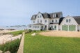 Large white coastal house with black roof shingles, multiple dormer windows, and a two-car garage with light green doors. The house is situated on a well-maintained green lawn next to a rocky shoreline with a view of the ocean and small islands in the distance. There are Adirondack chairs on a patio near the water.