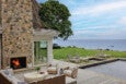 Outdoor patio area with wicker seating and cushions around a round coffee table, a stone fireplace with a fire burning, a wooden deck leading to a pool, several lounge chairs on a grassy area, and a large tree overlooking a calm body of water under a partly cloudy sky.