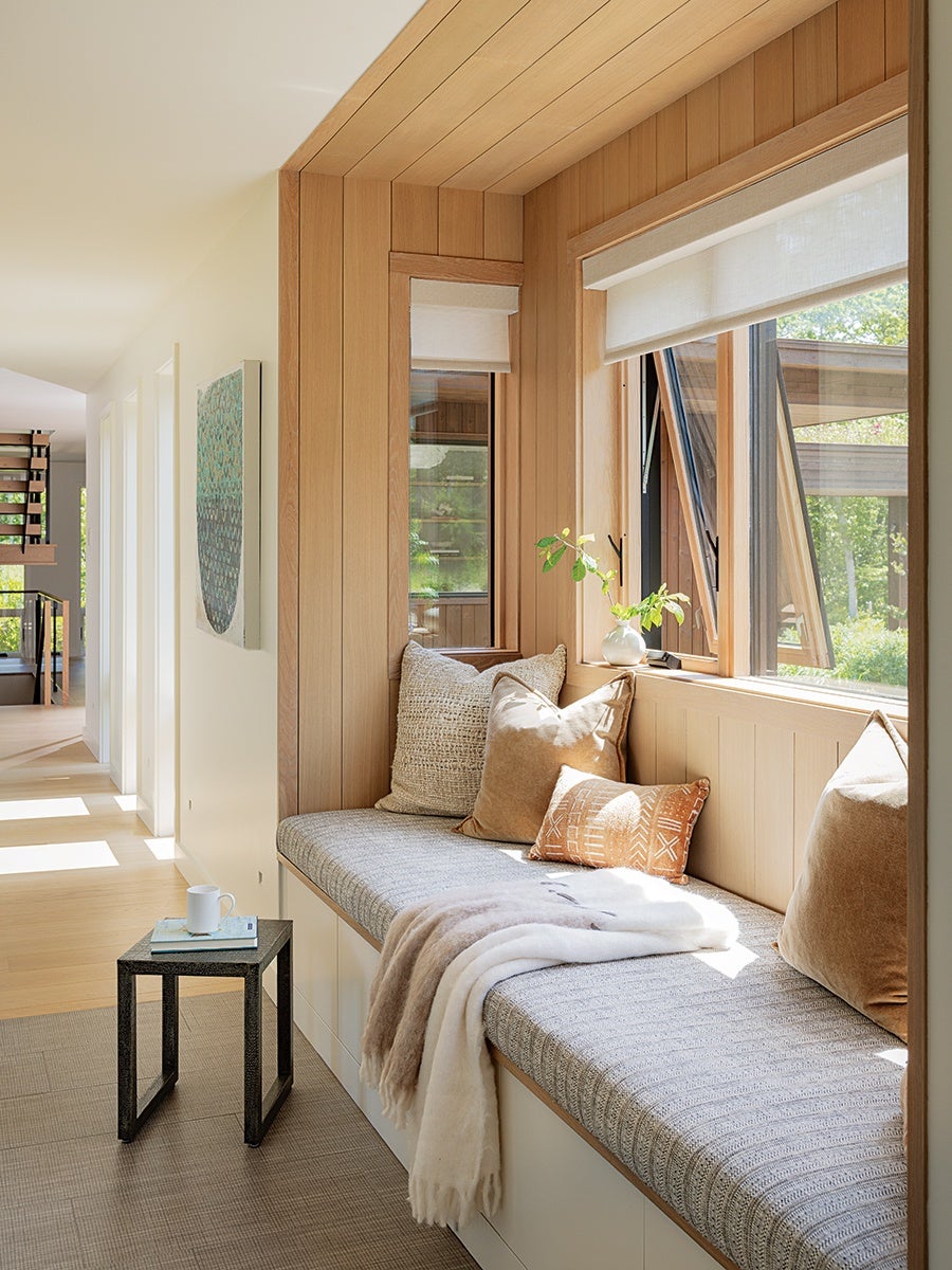 A cozy window seat with a gray textured cushion, adorned with various throw pillows in neutral tones and a small patterned orange pillow. A soft beige and white throw blanket is draped over the seat. The surrounding walls and ceiling are clad in light wood paneling. A small black side table next to the seat holds a white mug and a book. The window above the seat is open, letting in natural light and a view of greenery outside.