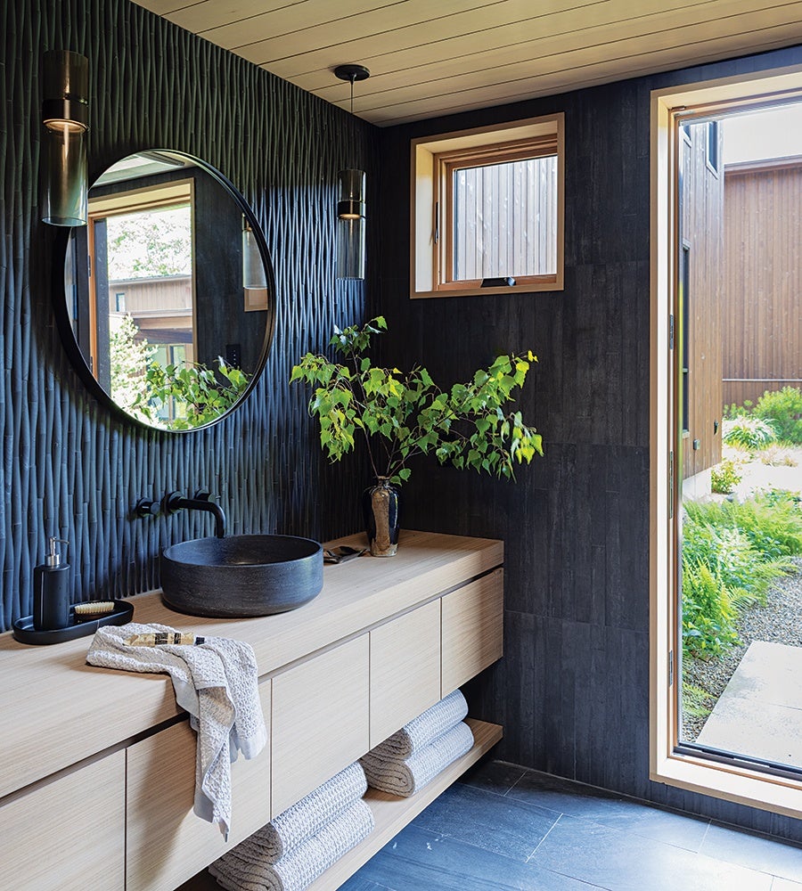 Modern bathroom with a textured dark wall featuring a large round mirror and two cylindrical pendant lights. A light wood floating vanity with a black stone vessel sink and black faucet sits below the mirror. A vase with green leafy branches adds a natural touch. Rolled towels are neatly stored on an open shelf beneath the vanity. A large window and a door with wooden frames open to a garden outside.