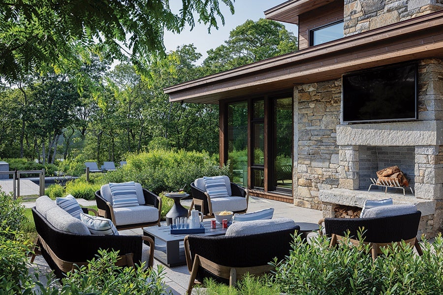 Outdoor patio seating area with cushioned chairs and a sofa arranged around a low coffee table. The patio is adjacent to a stone wall with a built-in fireplace and a mounted flat-screen TV above it. The setting is surrounded by greenery and trees, creating a serene and inviting atmosphere.