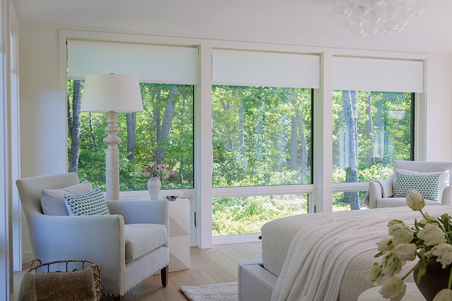 A bright bedroom featuring large windows with white roller shades partially down, allowing a view of lush green trees outside. The room includes two light gray upholstered armchairs with patterned throw pillows, a white bedside table with a vase of flowers, a tall white floor lamp, and a bed with a textured white blanket. A bouquet of white flowers is visible in the foreground. The flooring is light wood, and the overall decor is light and airy.