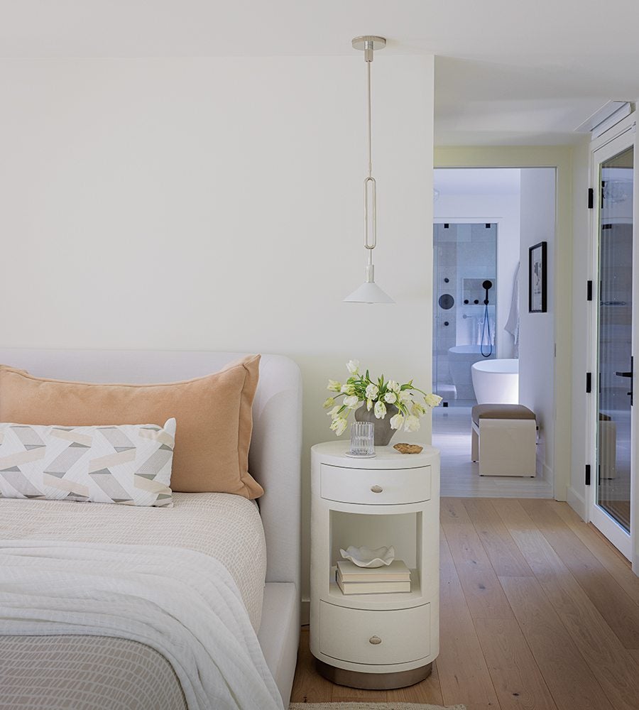 White bedroom with a beige bed featuring a large tan pillow and a geometric-patterned lumbar pillow. Next to the bed is a white round nightstand with two drawers and an open shelf holding books and a decorative bowl. On top of the nightstand is a vase of white flowers, a glass, and a small item. A white pendant light hangs above the nightstand. The room has light wood flooring and a doorway leading to a bathroom with a white freestanding bathtub and a bench.