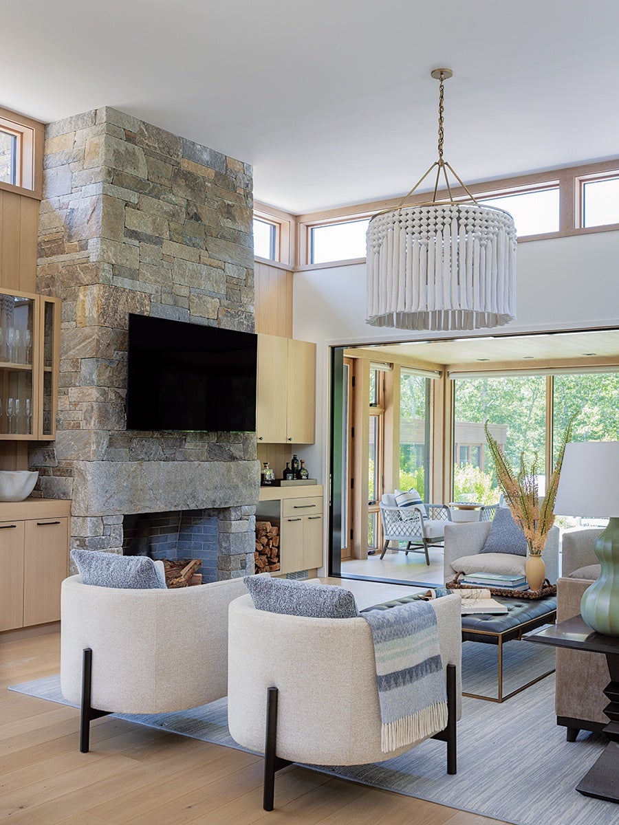 Modern living room with a stone fireplace and a mounted flat-screen TV. Two cream-colored armchairs with gray cushions and a blue-striped throw blanket face the fireplace. A large white fringed chandelier hangs from the ceiling. The room features light wood flooring and cabinetry, with large windows and sliding glass doors opening to a sunroom furnished with wicker chairs and a coffee table. A green vase with dried plants and a table lamp are on a side table next to a gray sofa.