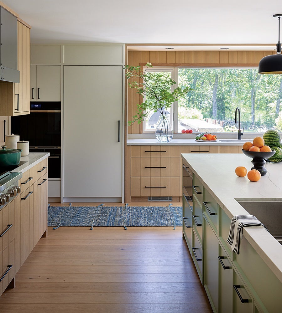 Modern kitchen with light wood cabinetry and a large island featuring a white countertop and built-in sink. A black bowl filled with oranges and loose oranges sit on the island, which also has a striped towel hanging over the edge. The back wall has a large window overlooking greenery, a vase with leafy branches on the counter, and a plate of assorted vegetables. The floor is light wood with a blue woven rug near the refrigerator and oven area. Black hardware and a black pendant light add contrast to the light, natural tones.
