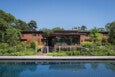 Modern wooden house with large glass windows, surrounded by lush greenery, and a clear blue swimming pool in the foreground under a bright blue sky.