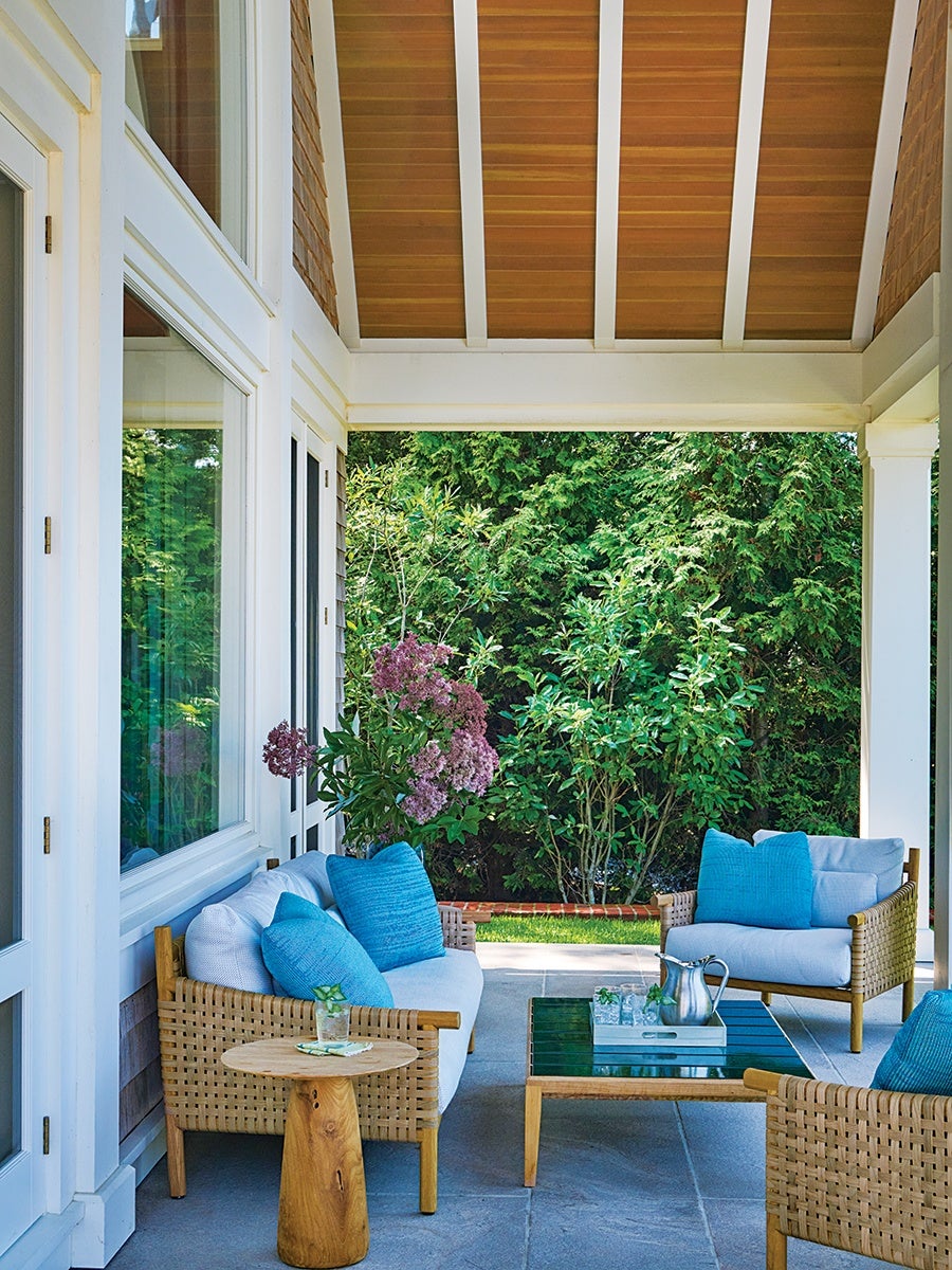 Covered outdoor patio with wicker furniture featuring white cushions and blue pillows, a wooden side table with a glass of water, and a glass-top coffee table holding a silver pitcher and glasses, surrounded by lush green plants and trees. The ceiling is wooden with white beams, and the floor is stone.