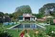A backyard scene featuring a rectangular swimming pool surrounded by a concrete border. On the left side of the pool, there is a seating area with light blue outdoor furniture including chairs and a sofa. On the right side, there are four light blue lounge chairs under two large white umbrellas. The pool area is bordered by well-maintained green grass and colorful flower beds in the foreground. In the background, there is a small house with a wooden pergola over a patio, surrounded by trees and brick walls. The sky is clear and blue.