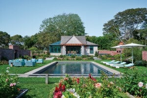 A backyard scene featuring a rectangular swimming pool surrounded by a concrete border. On the left side of the pool, there is a seating area with light blue outdoor furniture including chairs and a sofa. On the right side, there are four light blue lounge chairs under two large white umbrellas. The pool area is bordered by well-maintained green grass and colorful flower beds in the foreground. In the background, there is a small house with a wooden pergola over a patio, surrounded by trees and brick walls. The sky is clear and blue.