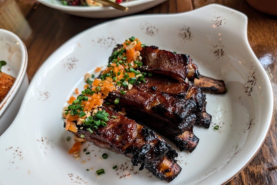 Grilled lamb ribs with a charred exterior, topped with finely chopped green herbs and small orange vegetable pieces, served in a white bowl with a speckled pattern.