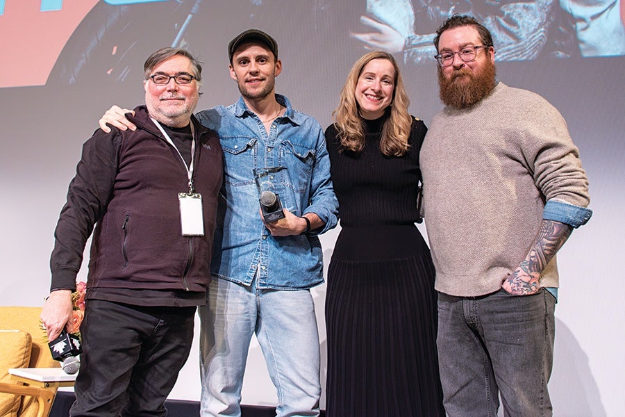 Four people stand together smiling. From left to right: a man with glasses, gray hair, and a beard wearing a dark zip-up jacket and black pants; a man in a denim jacket and light jeans holding a microphone and an award; a woman with long blonde hair wearing a black long-sleeve dress; and a man with glasses, a beard, and tattoos on his right arm wearing a beige sweater with rolled-up sleeves and dark jeans. They are standing indoors against a plain background with part of a screen visible above.