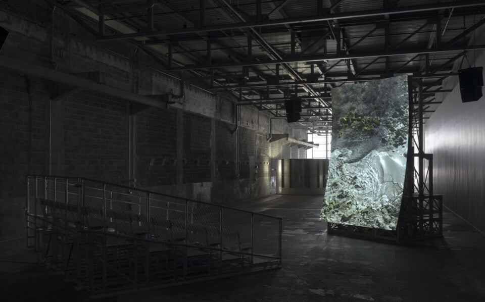 A large, tilted screen displaying an aerial view of a rocky shoreline with water and some greenery is set up inside a dimly lit industrial space with concrete walls and exposed ceiling beams. In front of the screen, there is a metal bleacher seating structure with a chain-link fence railing. The space has a utilitarian, warehouse-like atmosphere.