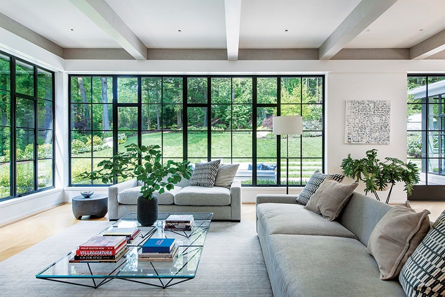 Modern living room with large black-framed windows overlooking a green garden. The room features two light gray sofas with patterned and solid pillows, a glass coffee table with books and a black vase holding green leafy branches, a floor lamp, and a small round side table. Light wood flooring and white walls with exposed ceiling beams complete the space.