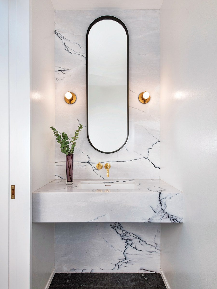 A modern bathroom vanity with white marble featuring dark veining. It has a rectangular sink integrated into the marble countertop and backsplash. Above the sink is a tall, narrow, oval mirror with a black frame. On either side of the mirror are round gold wall sconces with white bulbs. A slim purple vase with green leafy branches is placed on the left side of the countertop. The walls surrounding the vanity are plain white, and the floor is dark.