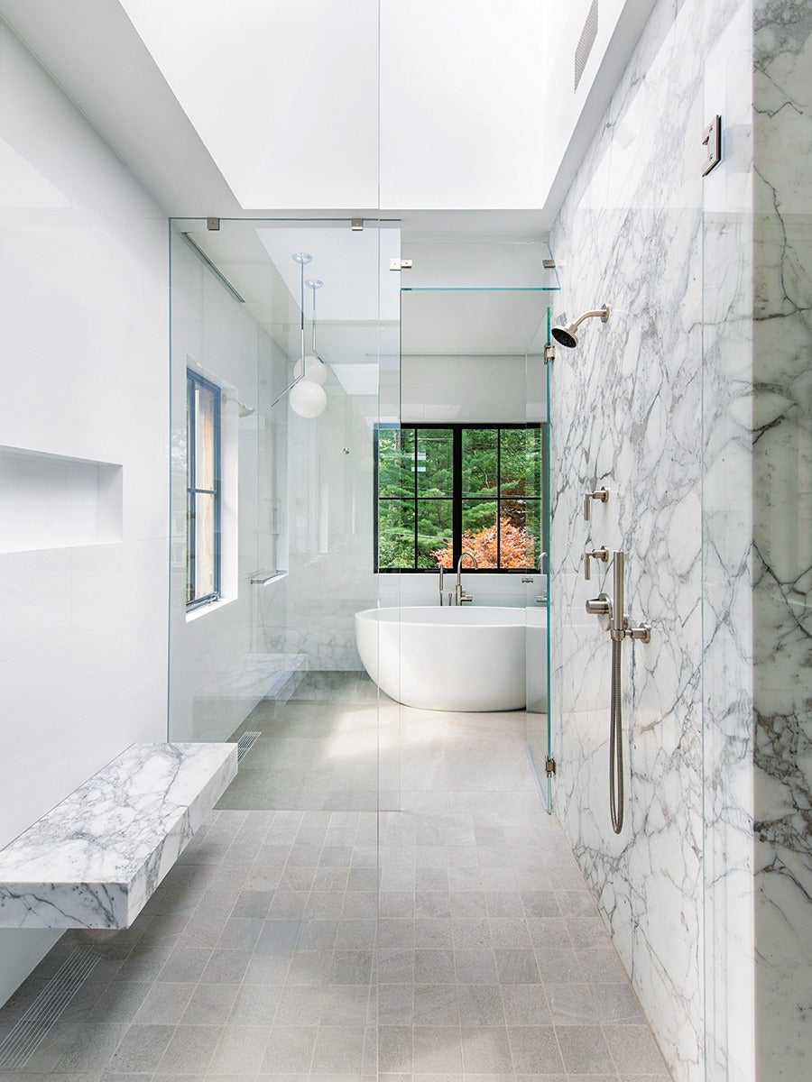 Modern bathroom with a large walk-in shower featuring marble walls, a built-in marble bench, and a glass partition. In the background, a freestanding white bathtub is positioned near a window with black frames, offering a view of green and orange foliage outside. The floor is tiled in light gray, and the space is illuminated by natural light from a skylight above.