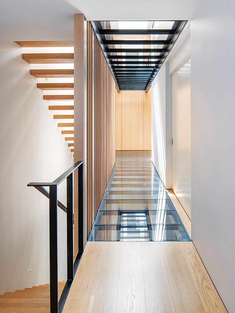A modern interior hallway features a glass floor section with wooden flooring on either side. The left side has vertical wooden slats and a black metal railing next to a staircase with floating wooden steps. The ceiling above the glass floor has black metal beams with glass panels, allowing natural light to filter through. The walls are white, and the far end of the hallway has a light wood panel wall.