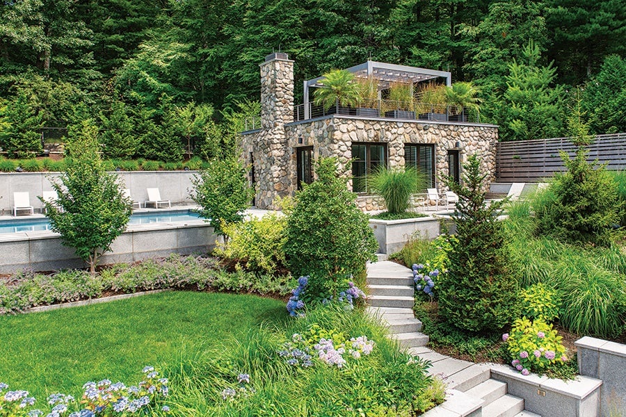 Stone house with a rooftop garden featuring palm plants, surrounded by lush greenery and a well-maintained lawn. Concrete steps lead up to the house, and there is a swimming pool with white lounge chairs on the left side. Tall trees and dense forest form the background.