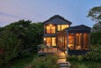 Modern two-story house with large windows and a wooden deck, illuminated warmly from inside during dusk. The house features a screened porch on the right side and is surrounded by lush greenery and garden plants. Steps with built-in lighting lead up to the deck.