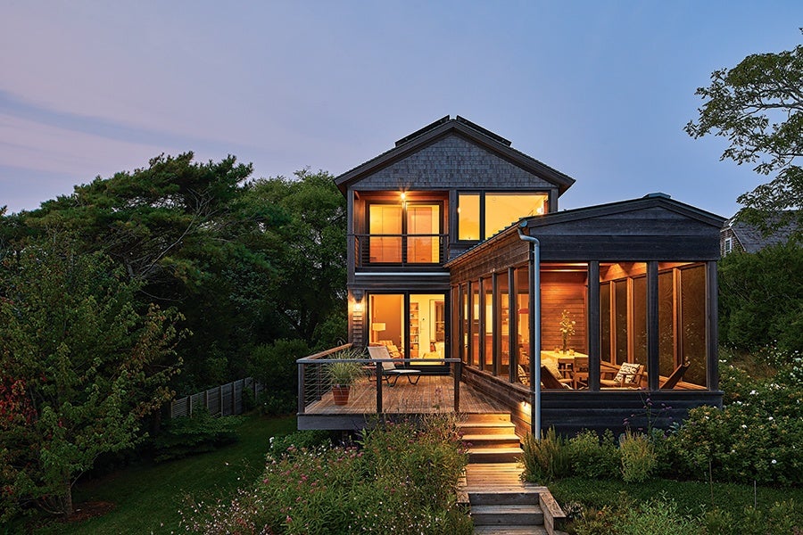 Modern two-story house with large windows and a wooden deck, illuminated warmly from inside during dusk. The house features a screened porch on the right side and is surrounded by lush greenery and garden plants. Steps with built-in lighting lead up to the deck.
